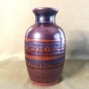 May include: A ceramic vase with a glossy finish, featuring a deep purple and dark brown color palette. The body is decorated with two horizontal bands of textured, burnt orange glaze that have a drip-like pattern along the edges.
