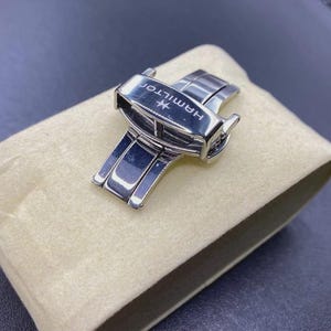 Peut inclure: Une boucle déployante de montre en acier inoxydable argenté avec une finition polie et le nom de la marque Hamilton gravé sur la surface supérieure. Le mécanisme est présenté en position fermée, montrant sa structure métallique et sa marque.