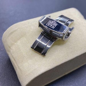 Può includere: Una chiusura deployante per orologio in acciaio inossidabile color argento con finitura lucida e una piastra centrale rettangolare nera con il logo bianco TAG HEUER. Il meccanismo presenta un design pieghevole incernierato con pulsanti laterali.