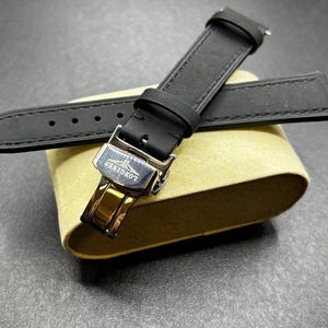 Könnte beinhalten: Ein schwarzes Uhrenarmband mit einer silberfarbenen Metall-Faltschließe, auf deren Oberfläche das Longines-Logo und der Markenname eingraviert sind. Das Armband hat sichtbare Nähte an den Kanten und eine passende schwarze Schlaufe.