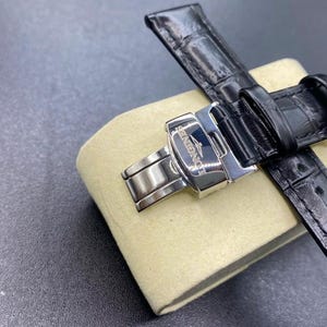 May include: A black leather watch strap with a glossy alligator-embossed texture and a polished silver-tone stainless steel deployment clasp. The metal clasp features the engraved Longines brand name and logo on its surface.