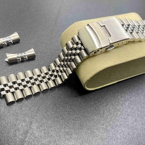 May include: A silver-toned stainless steel jubilee watch bracelet with a fold-over clasp and two curved end links. The metal watch band features a multi-link design with a brushed and polished finish, resting on a beige display cushion.