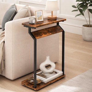 May include: A C-shaped side table featuring a rustic brown wood-grain finish on two rectangular shelves supported by a black metal frame. The top shelf holds a lamp, a candle, and a picture frame, while the bottom shelf holds a white vase and books.