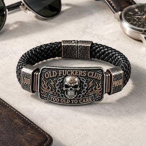 Könnte beinhalten: Ein schwarzes geflochtenes Armband mit einem rechteckigen Metallmittelstück, das einen Totenkopf, Ketten und Flammen zeigt. Der Text lautet OLD FUCKERS CLUB und TOO OLD TO CARE, mit LOUD und PROUD auf den seitlichen Metallakzenten in dunkler Optik.