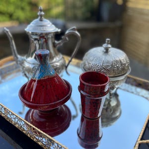 May include: A rectangular mirrored serving tray with gold-toned filigree handles holds a silver teapot, a silver lidded bowl with intricate floral engravings, a small red and blue ceramic tagine, and a matching red ceramic cup with a speckled finish.