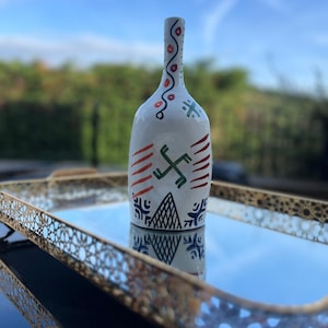 Puede incluir: Una botella de cerámica blanca con cuello estrecho, decorada con motivos pintados a mano que incluyen un símbolo geométrico verde central, líneas horizontales rojas y patrones triangulares negros. La botella presenta un cuello cilíndrico y delgado con una línea roja.