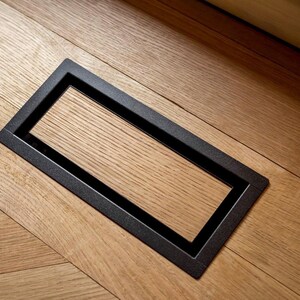 May include: A rectangular black metal floor vent cover set flush into light brown oak wood flooring. The vent features a clean, minimalist frame surrounding a matching wood insert that aligns with the grain of the surrounding floorboards.