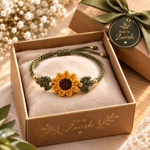 May include: A macrame bracelet featuring a central crocheted sunflower with yellow petals and a brown center, flanked by green leaves and small gold beads. The bracelet has a green cord band and is presented in a brown gift box with the text Made by Zarish.