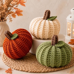 May include: Three crocheted pumpkins in rust orange, cream, and sage green. Each pumpkin features a textured ribbed pattern, a brown crocheted stem, and a small green leaf attached at the top. The pumpkins are arranged in a cluster.