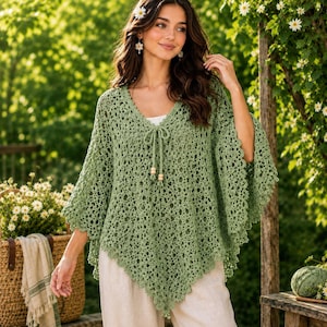 May include: A sage green crochet poncho featuring an intricate openwork pattern, a V-neckline with a drawstring closure, and two wooden beads at the ends of the ties. The garment has a pointed hemline and wide, loose sleeves with a scalloped edge.