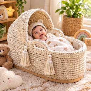 May include: A cream-colored crocheted baby bassinet with a hood, featuring two decorative tassels hanging from the front. Inside, a baby wears a matching knit hat and is covered by a white blanket patterned with small brown teddy bear faces.