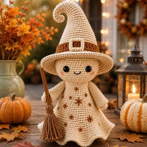May include: A cream-colored crochet doll wearing a pointed witch hat with a brown band and buckle. The doll has black bead eyes, a small smile, and pink cheeks. The dress features embroidered brown star patterns, and the doll holds a small broom.