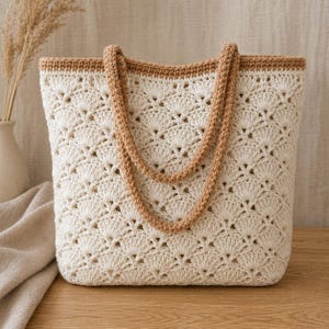 Peut inclure: Un sac cabas en crochet avec un corps blanc cassé présentant un motif de points en éventail répétitif. Le sac possède une bordure marron clair unie sur le haut et deux anses en crochet assorties. La structure est entièrement textile.