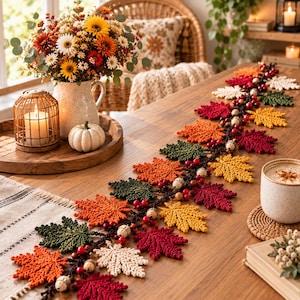 Può includere: Una ghirlanda decorativa con foglie d'acero all'uncinetto nei toni del rosso, arancione, giallo, verde e crema, intervallate da piccole bacche rosse e perline di legno, disposte in una lunga linea sinuosa su un tavolo di legno.