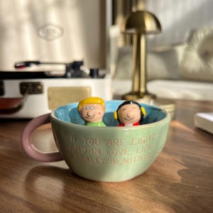May include: A light green ceramic mug with a pink handle and a light blue interior. Inside the mug are two small character figurines. The exterior features the embossed text: IF YOU ARE EIGHT AND IN LOVE, LIFE IS REALLY BEAUTIFUL with a small heart symbol.