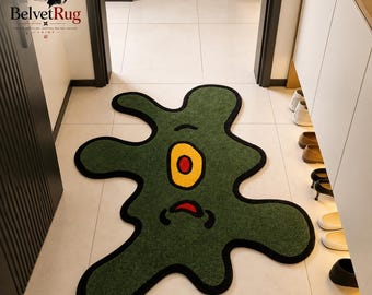 Pode incluir: Um tapete de formato irregular semelhante a uma ameba na cor verde-oliva escuro. O tapete apresenta um olho central oval amarelo com uma pupila vermelha e uma pequena boca preta curva abaixo. Todo o formato é contornado por uma borda preta.
