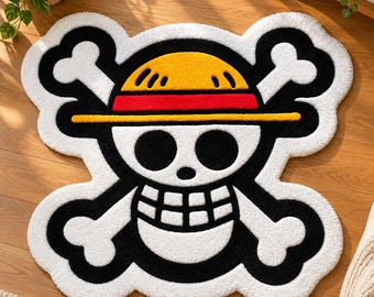 Pode incluir: Um tapete tufado com o formato do emblema de caveira e ossos cruzados de One Piece. O design apresenta uma caveira branca com órbitas e dentes pretos, usando um chapéu de palha amarelo com uma faixa vermelha sobre um fundo preto com ossos.