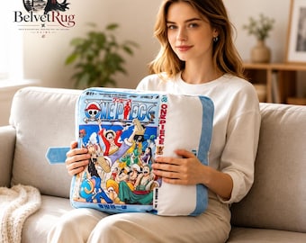 Pode incluir: Uma almofada retangular com uma impressão gráfica colorida da capa do mangá One Piece, incluindo o título e várias ilustrações de personagens. A almofada tem um painel lateral branco e debrum azul-claro ao longo das bordas.