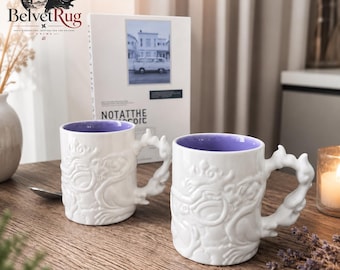 Pode incluir: Duas canecas de cerâmica branca com um design intrincado de dragão em relevo no exterior e um interior roxo sólido. Cada caneca tem uma alça de formato único que imita a aparência de fumaça ou chamas rodopiantes para uma aderência.
