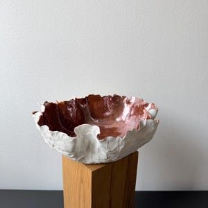 Op de afbeelding: Een keramische kom met een golvende, organische rand. De buitenkant van de kom is matwit met een textuur die op een blad lijkt, terwijl de binnenkant een gladde, glanzende metallic koperen afwerking heeft die het licht op het oppervlak weerkaatst.