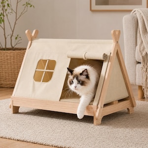 May include: A beige canvas pet tent with a light-colored wooden frame, featuring a square window with a cross-bar design and a roll-up front entrance. A fluffy cat with brown and white markings is stepping out of the tent opening.