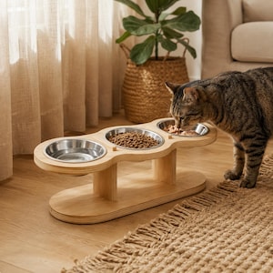 Puede incluir: Un comedero elevado de madera clara con tres aberturas circulares que sostienen cuencos extraíbles de acero inoxidable. El cuenco central contiene pienso seco, mientras que el de la derecha contiene comida húmeda. Un gato atigrado come.