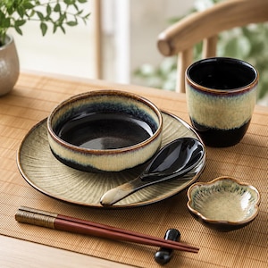 May include: A ceramic dinnerware set featuring a round plate, a bowl, a cup, a spoon, a small flower-shaped dish, and a pair of chopsticks. The pieces have a reactive glaze finish with a gradient of dark black, deep blue, and light beige tones.
