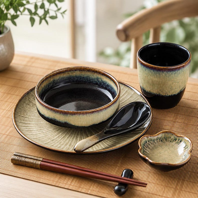 May include: A ceramic dinnerware set featuring a round plate, a bowl, a cup, a spoon, a small flower-shaped dish, and a pair of chopsticks. The pieces have a reactive glaze finish with a gradient of dark black, deep blue, and light beige tones.