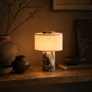 Może przedstawiać: Lampa stołowa z cylindryczną podstawą z białego i szarego żyłkowanego marmuru, zwieńczona okrągłym abażurem z tkaniny w kolorze złamanej bieli. Lampa emituje ciepłe, miękkie światło, a na górze znajduje się pasujący marmurowy element.