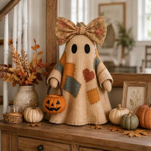 Puede incluir: Una figura de fantasma de tela color canela con un gran lazo de estampado floral en la cabeza y varios parches rectangulares en tonos azul, naranja y marrón. El fantasma sostiene un pequeño cubo de calabaza naranja con cara negra.