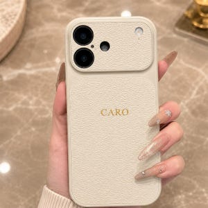 May include: A cream-colored smartphone case with a textured, leather-like finish. The back features a raised camera module with two circular lenses and a small flash. The name CARO is embossed in gold lettering in the center of the case.