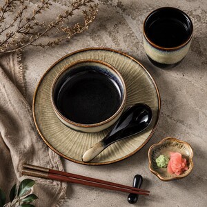 May include: A ceramic dinnerware set featuring a plate, a bowl, a cup, a spoon, and chopsticks. The pieces have a reactive glaze with a dark black centre transitioning to a textured, light beige rim. A small scalloped dish holds ginger and wasabi.