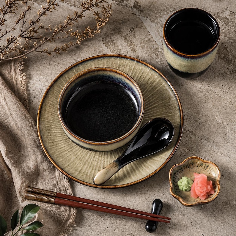 May include: A ceramic dinnerware set featuring a plate, a bowl, a cup, a spoon, and chopsticks. The pieces have a reactive glaze with a dark black centre transitioning to a textured, light beige rim. A small scalloped dish holds ginger and wasabi.