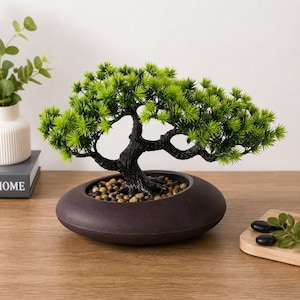 May include: An artificial bonsai tree with vibrant green needle-like foliage and a dark textured trunk. The tree is planted in a low, round, dark brown ceramic pot filled with small, smooth, multi-coloured decorative river stones.