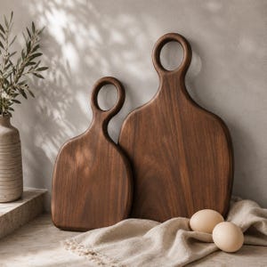 May include: Two dark brown wooden cutting boards with smooth, rounded edges and circular handles at the top. The boards feature a rich, natural wood grain pattern and are displayed in a vertical orientation, with one board slightly smaller than the other.