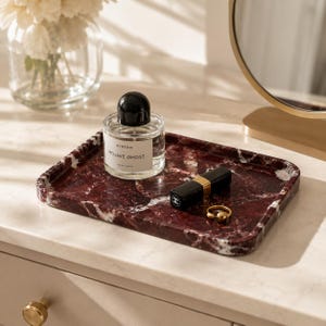May include: A rectangular dark red marble tray with white veining. Resting on the tray is a clear glass perfume bottle with a black cap labelled Byredo Mojave Ghost, a black lipstick tube with gold accents, and a small gold-toned metal ring.