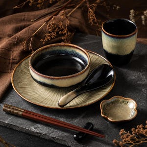 May include: A ceramic dinnerware set featuring a round plate with a textured beige sunburst pattern, a matching bowl with a dark interior, a glossy black spoon, a small flower-shaped dish, a cup with a beige and black gradient, and wooden chopsticks.