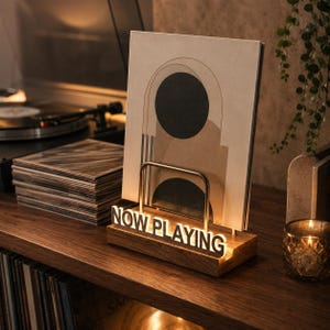 May include: A wooden vinyl record display stand featuring a metal arch and an illuminated sign that reads NOW PLAYING in white capital letters. The stand holds a square record sleeve with a beige, black, and brown abstract geometric design.