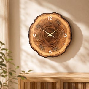 Può includere: Un orologio da parete rotondo realizzato con una fetta di legno naturale con anelli di crescita marrone scuro visibili e un bordo in corteccia grezza. Il quadrante mostra numeri bianchi alle posizioni 12, 3, 6 e 9 con lancette in metallo dorato.
