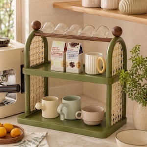 Puede incluir: Un estante de almacenamiento de dos niveles en color verde oliva con laterales arqueados que presentan paneles de rejilla clara. El estante superior sostiene cinco vasos de vidrio transparente y dos cajas etiquetadas White Chocolate Chunks y Semisweet Chocolate Chunks.