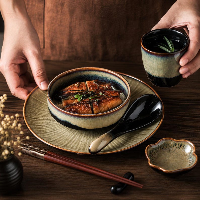 May include: A ceramic dinnerware set featuring a bowl, a plate, a spoon, a small cup, and chopsticks. The pieces have a reactive glaze with a gradient of dark brown, beige, and blue-green tones. The bowl contains grilled eel over white rice.