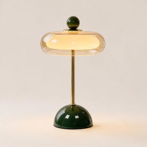 Puede incluir: Una lámpara de mesa con base en forma de cúpula verde oscuro y un remate esférico a juego sobre un vástago de metal dorado. La lámpara incluye una pantalla de cristal transparente tintado en ámbar que rodea un difusor de luz de cristal blanco.