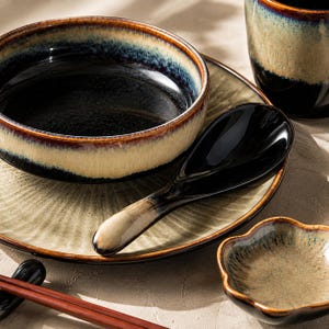 May include: A ceramic dinnerware set featuring a round bowl with a deep black interior and a cream-coloured exterior rim, a matching plate with a radial textured pattern, a black soup spoon, a small flower-shaped dish, and a pair of red chopsticks.