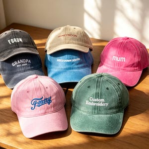 Op de afbeelding: Een collectie van zeven baseballpetten in diverse kleuren, waaronder antraciet, beige, blauw, roze en groen. Elke pet is voorzien van geborduurde tekst zoals FORUM, HONEYMOON, DISCO DOWN SOUTH, GRANDPA EST. 2024, mum, Forty en Custom Embroidery.
