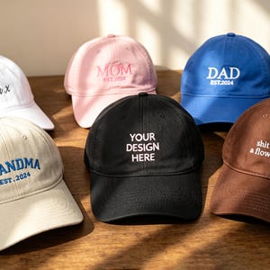 May include: A collection of six baseball caps in white, pink, blue, beige, black, and brown. Each cap features embroidered text including "X.IV.X", "MOM EST.2024", "DAD EST.2024", "GRANDMA EST.2024", "YOUR DESIGN HERE", and "shit makes a flower grow".