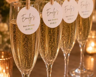 May include: Four clear glass champagne flutes filled with sparkling liquid, each featuring a round white paper tag attached to the rim with a small wooden clothespin. The tags are printed with the text Emily's BABY SHOWER 20.05.26.