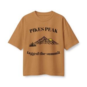 May include: A tan short-sleeve t-shirt featuring a graphic of a mountain range with a yellow sun, pine trees, and the black text PIKES PEAK above the mountain and tagged the summit below the mountain in a serif font on the front centre.