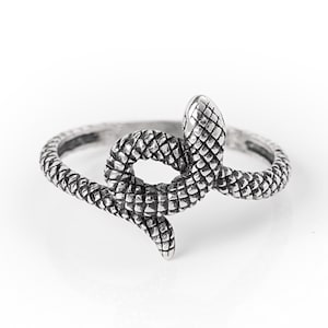 Peut inclure: Une bague argentée représentant un serpent enroulé. La surface métallique présente une finition oxydée sombre dans les zones en creux pour souligner le motif détaillé et texturé des écailles qui entoure tout l'anneau de la bague.