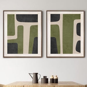 May include: Two framed abstract art prints with geometric shapes in shades of green, black, and beige. The prints are hung on a white wall above a wooden table with chairs.