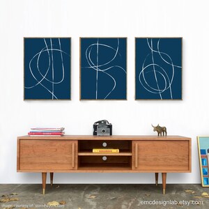 May include: Three framed abstract art prints with white lines on a dark blue background. The prints are hanging on a white wall above a wooden media console.